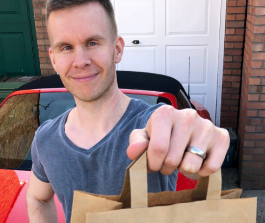 Volunteer Daniel standing in front of red car holding out food delivery bag.